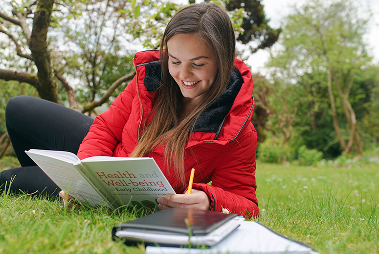 female looking at an early years textbook on grass holding pencil