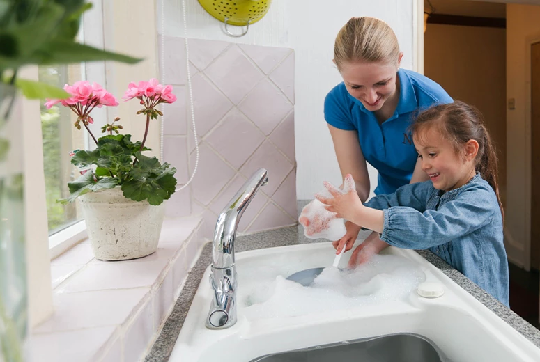 female student and young girl washing up in kitchen looking at bubbles