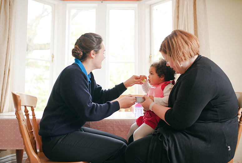 female nanny feeding little girl on mother's lap