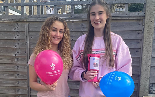 two females holding blue and pink balloons with a charity collection pot