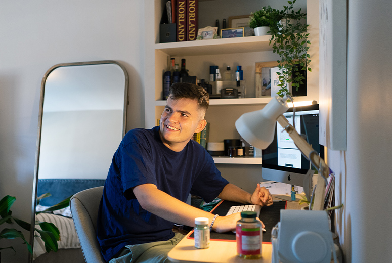 A male at a desk in his bedroom smiling at someone behind him