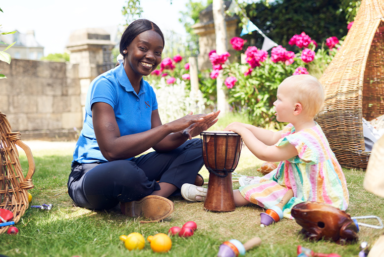 A female Norland Nanny student playing a drum with a child while looking at the camera smiling