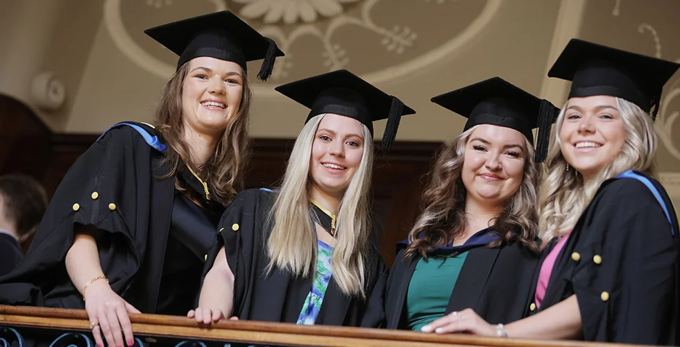 Four female graduates stood smiling looking over a balcony