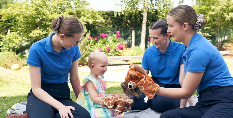 Three Norland Nanny students in their practical uniform with puppets playing with a girl who is smiling