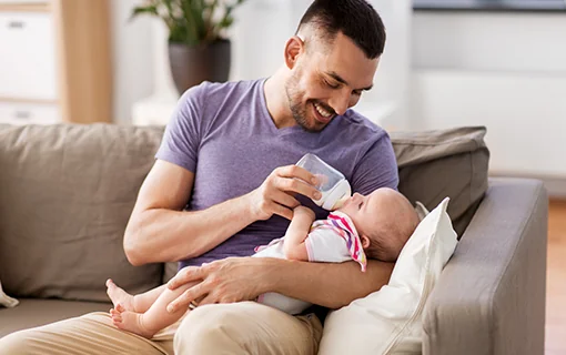 A male sat on a couch bottle feeding a baby