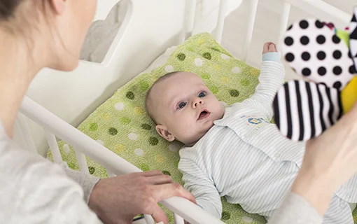 A baby led in a cot looking up at a toy that is being held by an adult