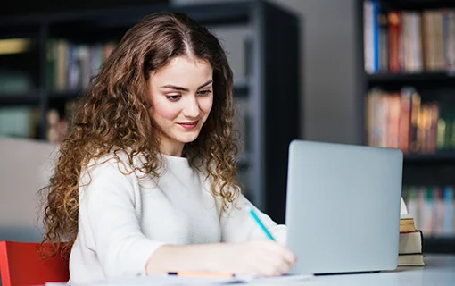 A lady sat at her laptop smiling, writing on a notepad