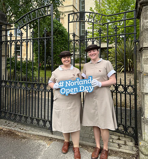 Norland students in their formal uniform outside the gates of the Upper Oldfield Park Campus in Bath holding a sign that says Norland Open Day