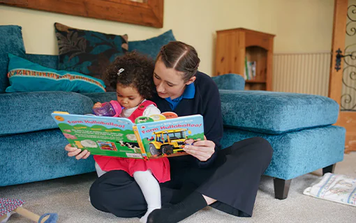 An adult sat reading a book to a child who is sat on her lap in a living room