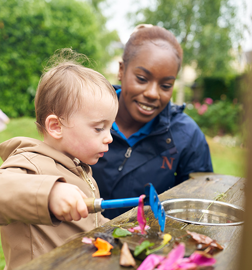 A Norland student outside with a child at a mud kitchen, and the child is holding some children's tools